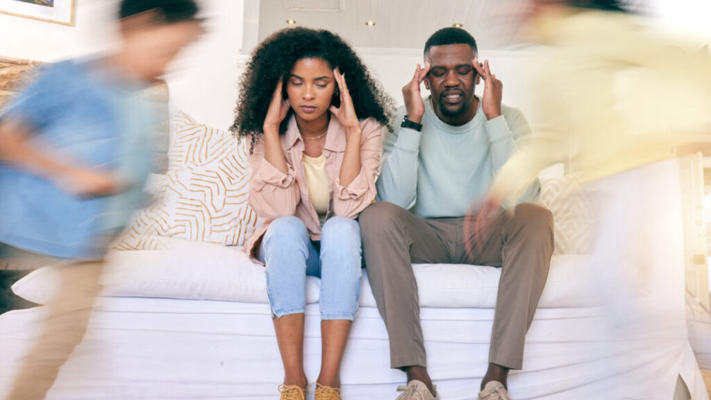 Tired parents on sofa with motion blur of energy as children run around house